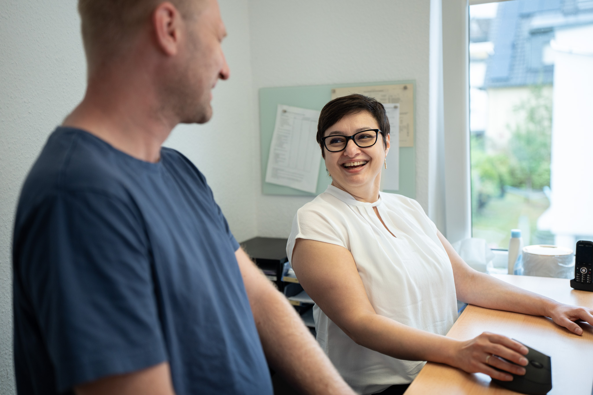 Lachende Frau mit Brille am Schreibtisch im Gespräch mit einem stehenden Kollegen im Büro.