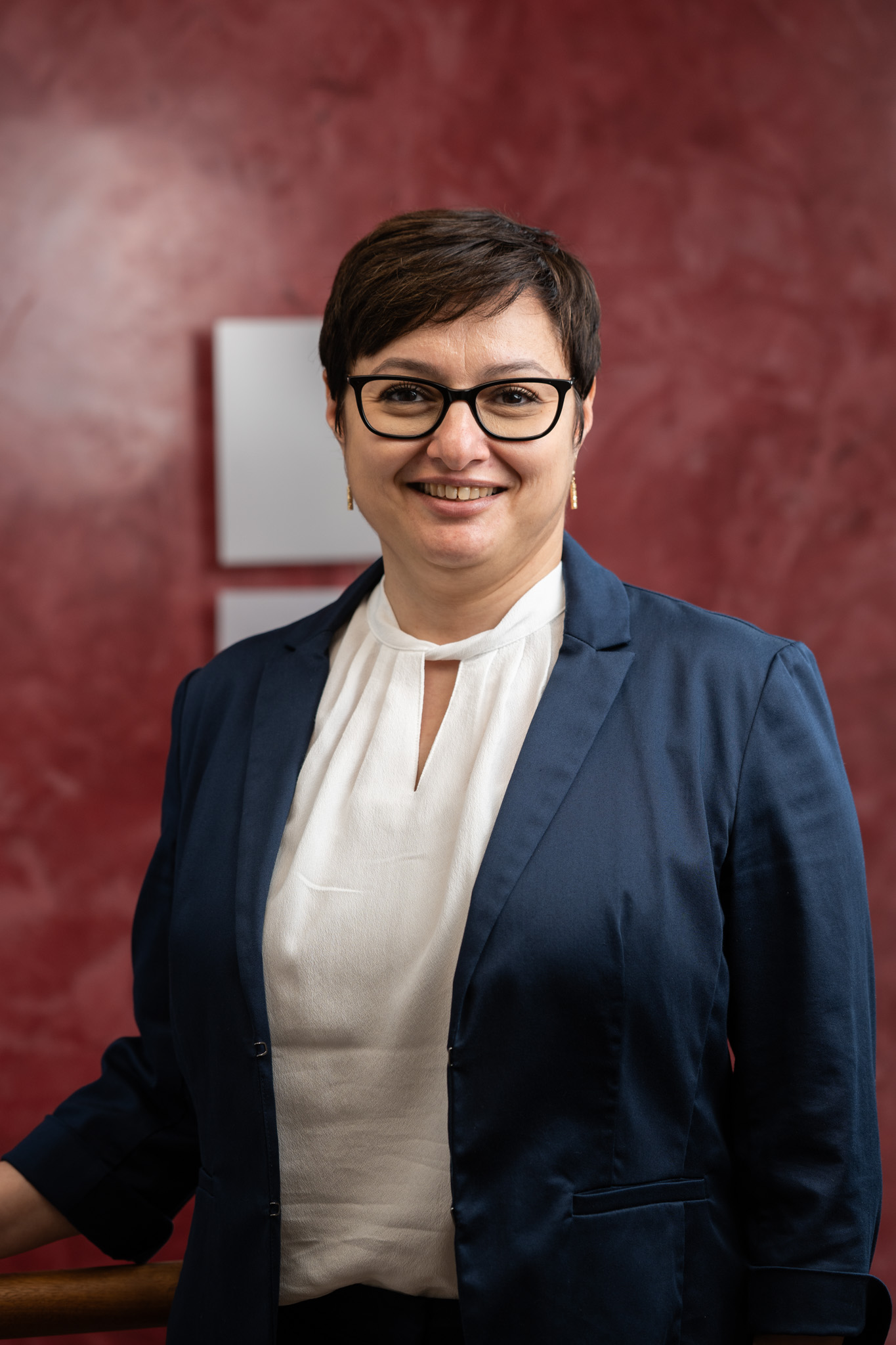 Porträt einer lächelnden Frau mit Brille, weißer Bluse und blauem Blazer vor rotem Hintergrund.