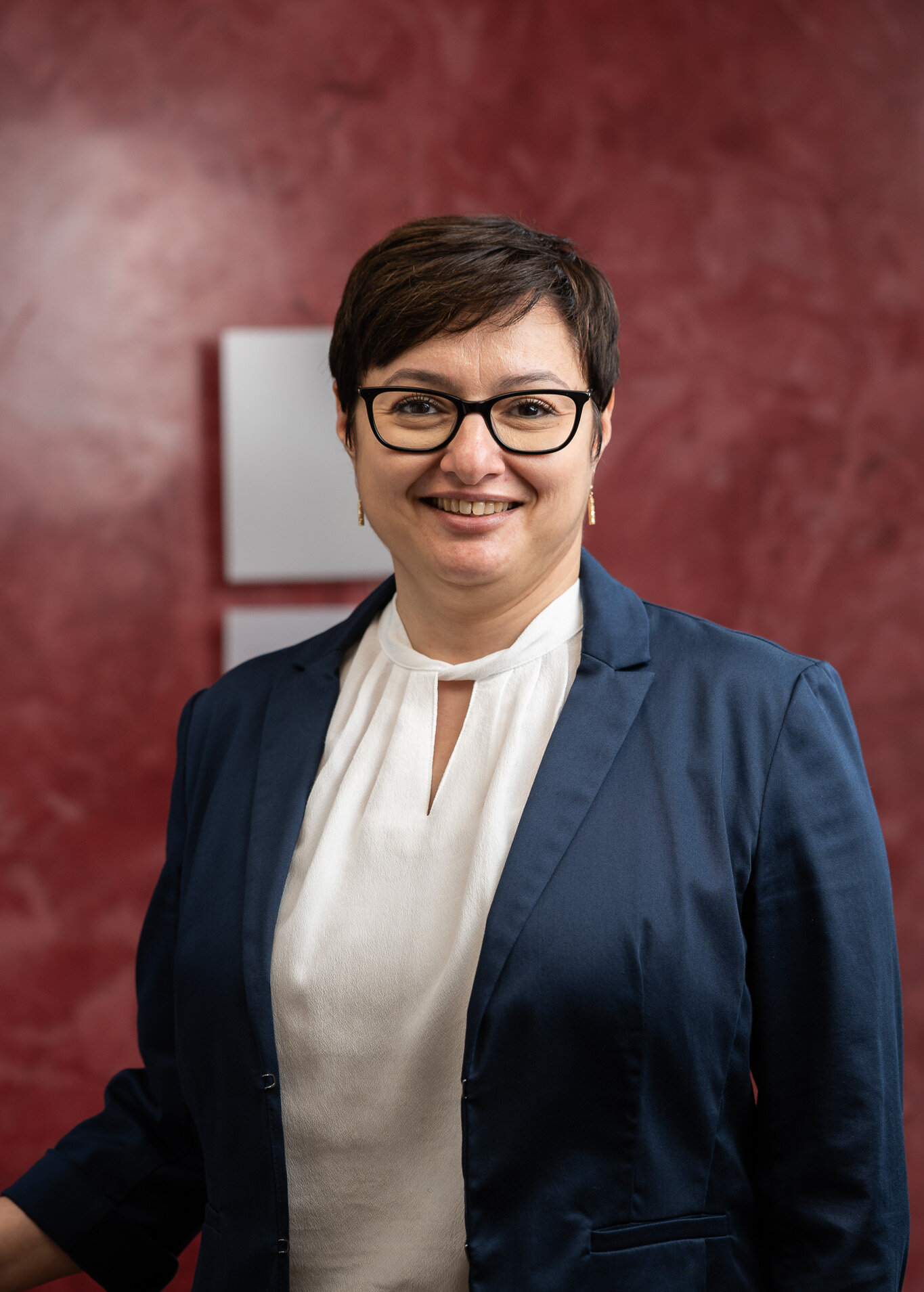 Porträt einer lächelnden Frau mit Brille, weißer Bluse und blauem Blazer vor rotem Hintergrund.