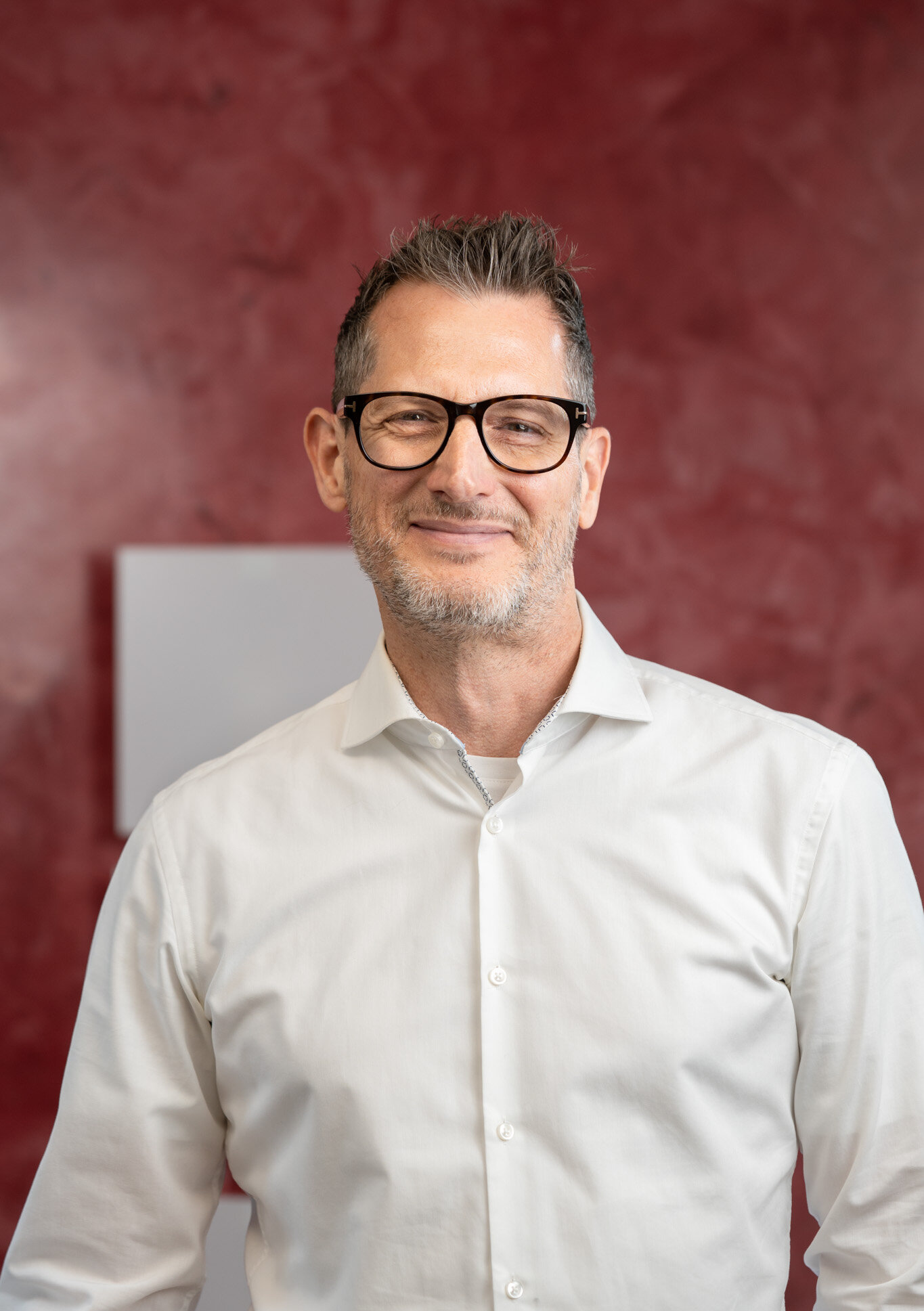 Porträt eines lächelnden Mannes mit Brille und weißem Hemd vor rotem Hintergrund.