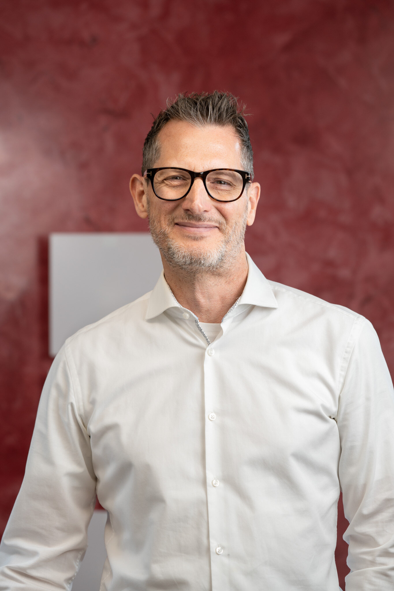 Porträt eines lächelnden Mannes mit Brille und weißem Hemd vor rotem Hintergrund.