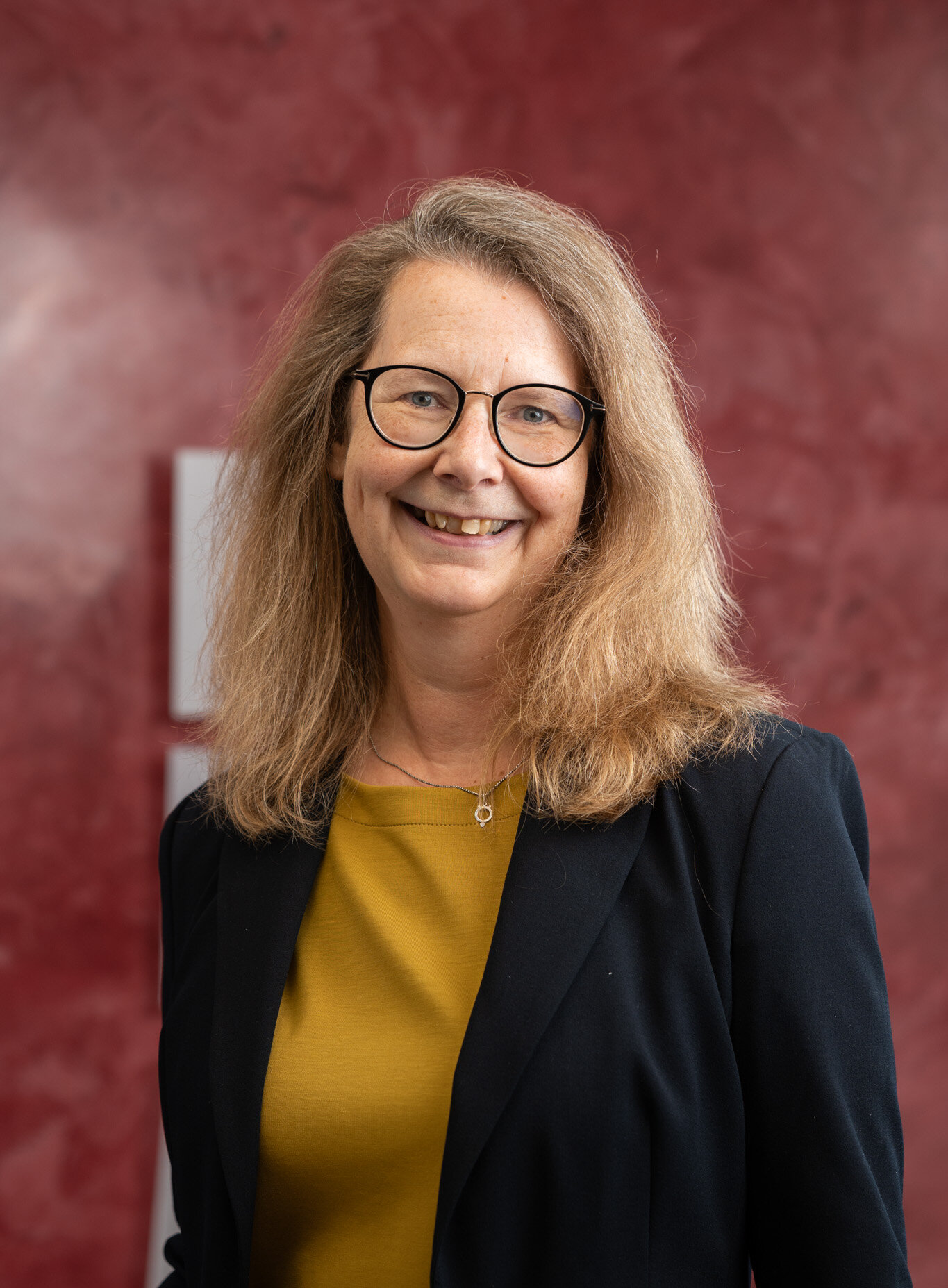 Porträt einer lächelnden Frau mit Brille, gelbem Top und dunklem Blazer vor rotem Hintergrund.